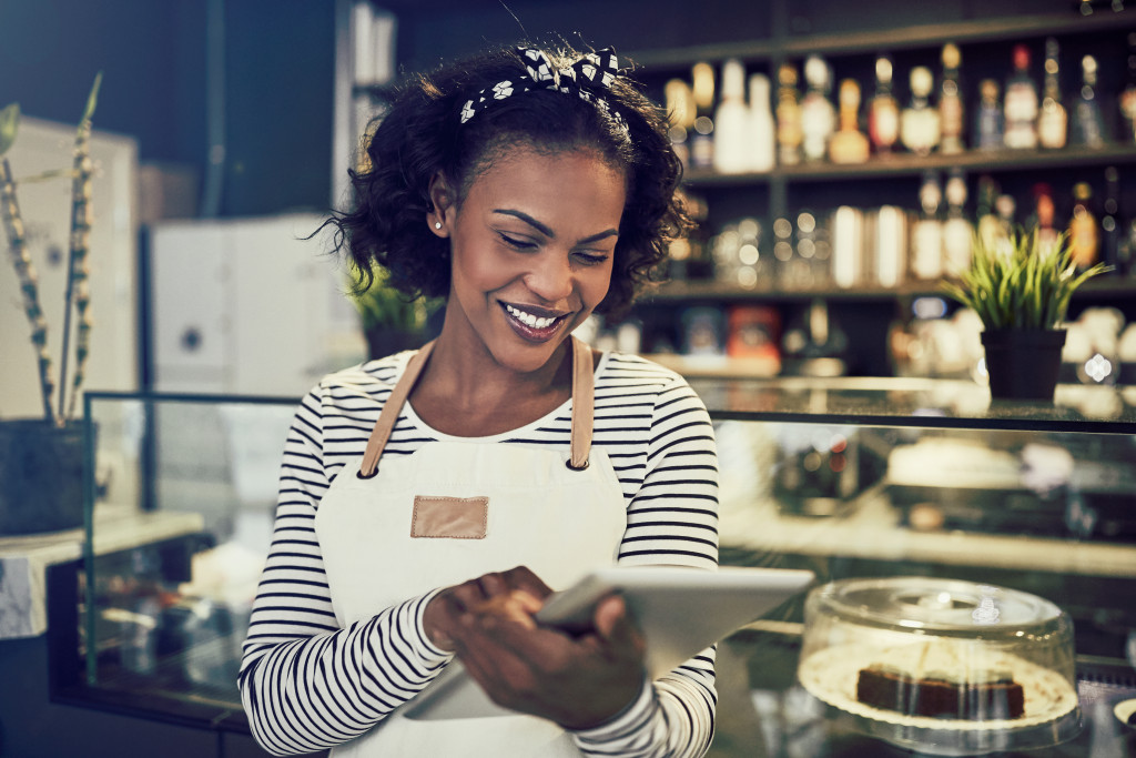 Young female business owner wearing a white apron while using a tablet in front of the cafe's counter.